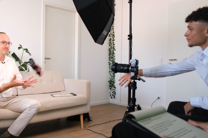 Interview-Aufbau vor Ort: eine Person sitzt sprechend auf einem Sofa, davor Kamera auf Stativ, Softbox und Teleprompter